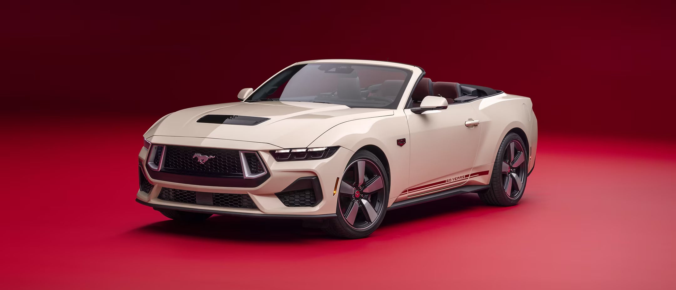 Front view of a convertible 2025 Ford Mustang in cream color against a bold red background.