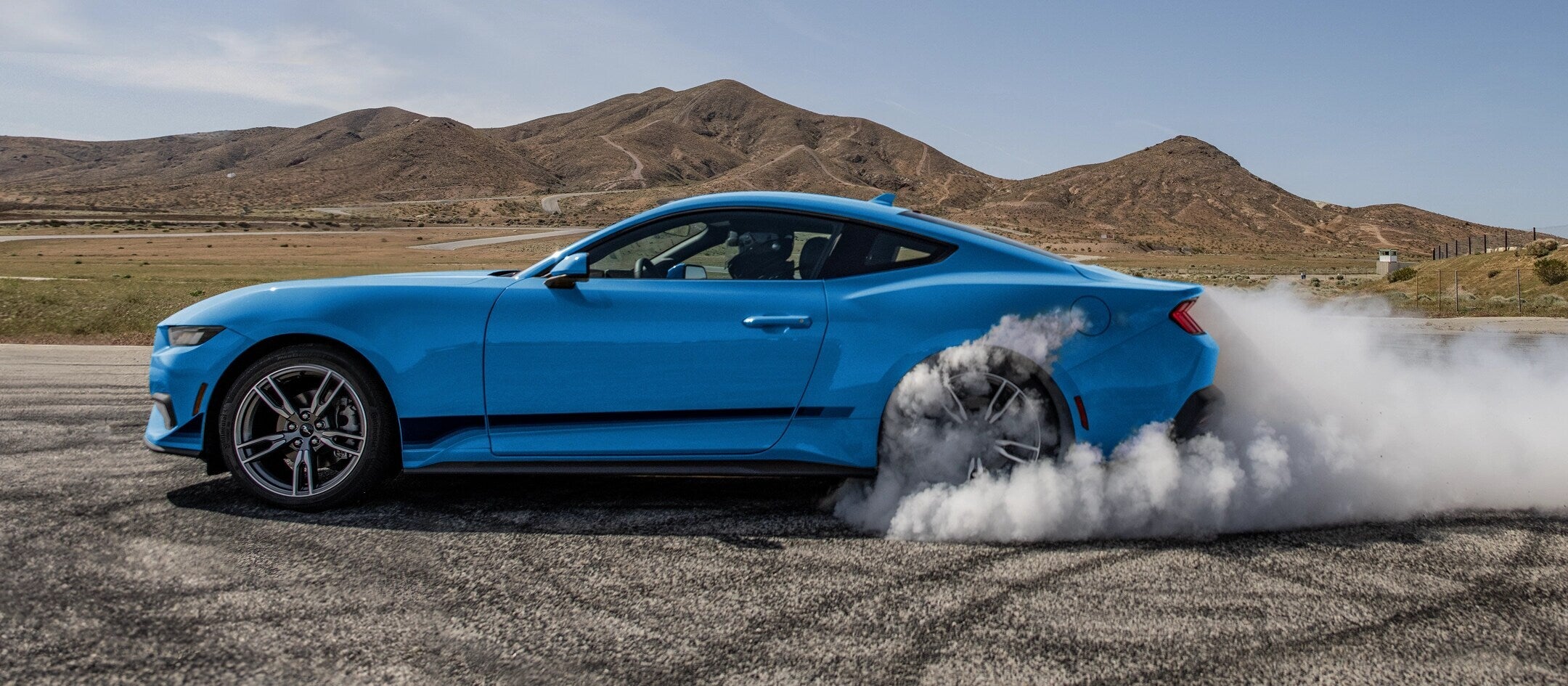 Side view of a blue 2025 Ford Mustang performing a burnout on a track, with smoke billowing from the rear tires.