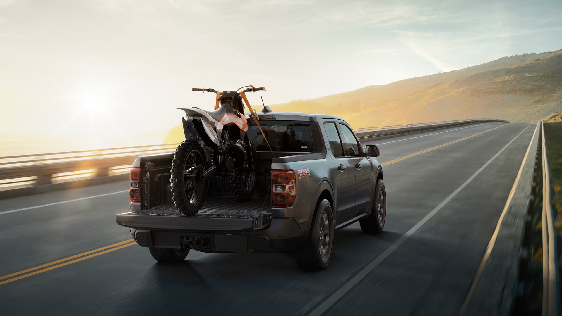 a 2026 Ford Maverick driving on a road with a dirt bike loaded inn the bed